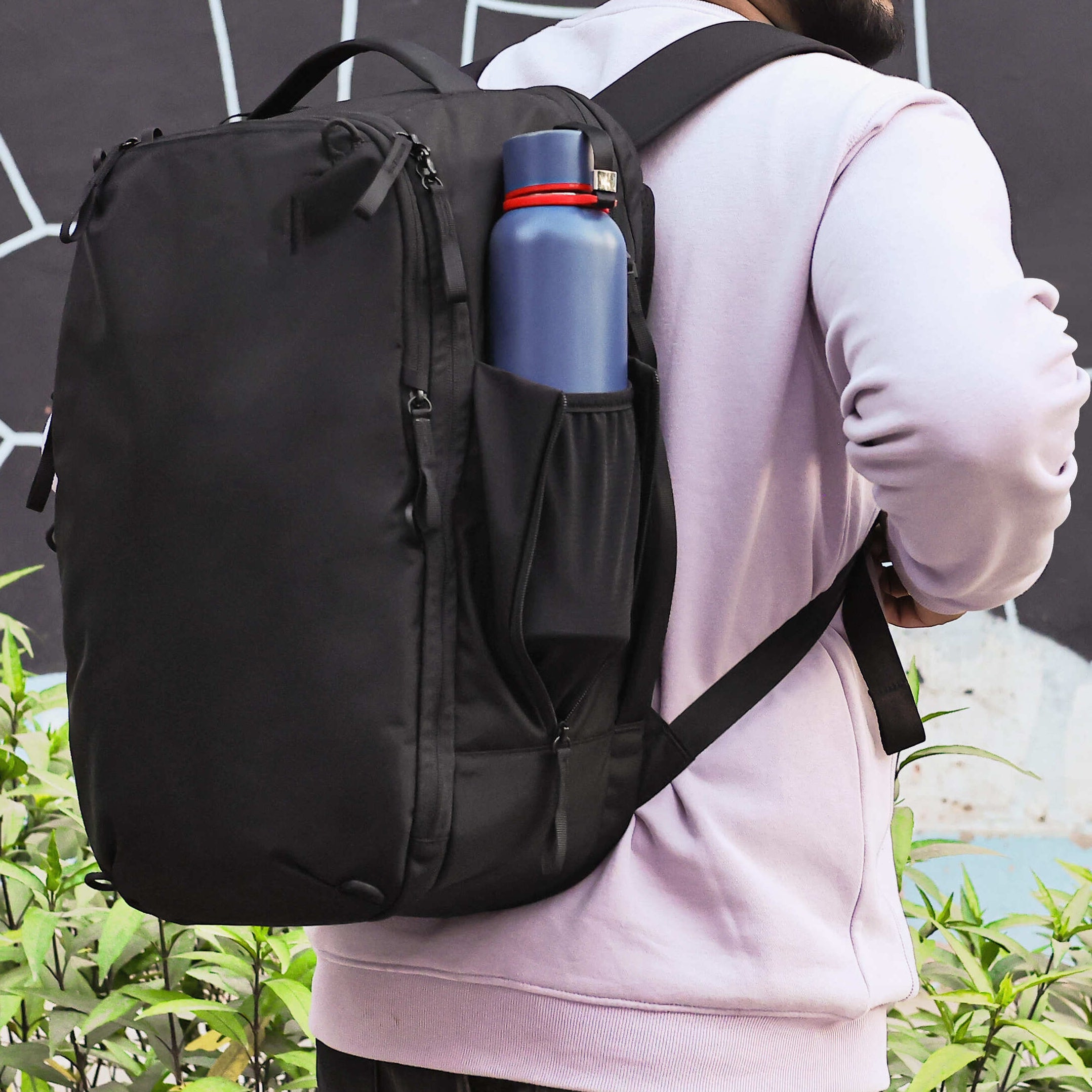 Man wearing a black backpack with a blue water bottle against a geometric-patterned wall.